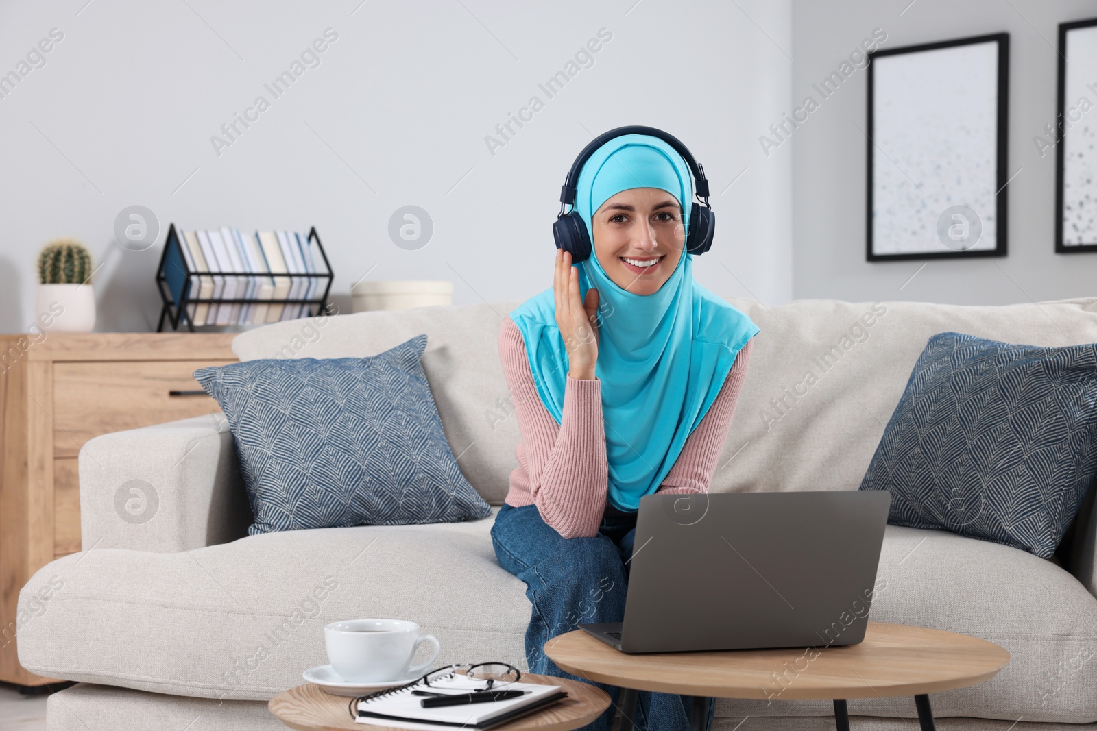 Photo of Muslim woman in headphones using laptop at wooden table in room. Space for text