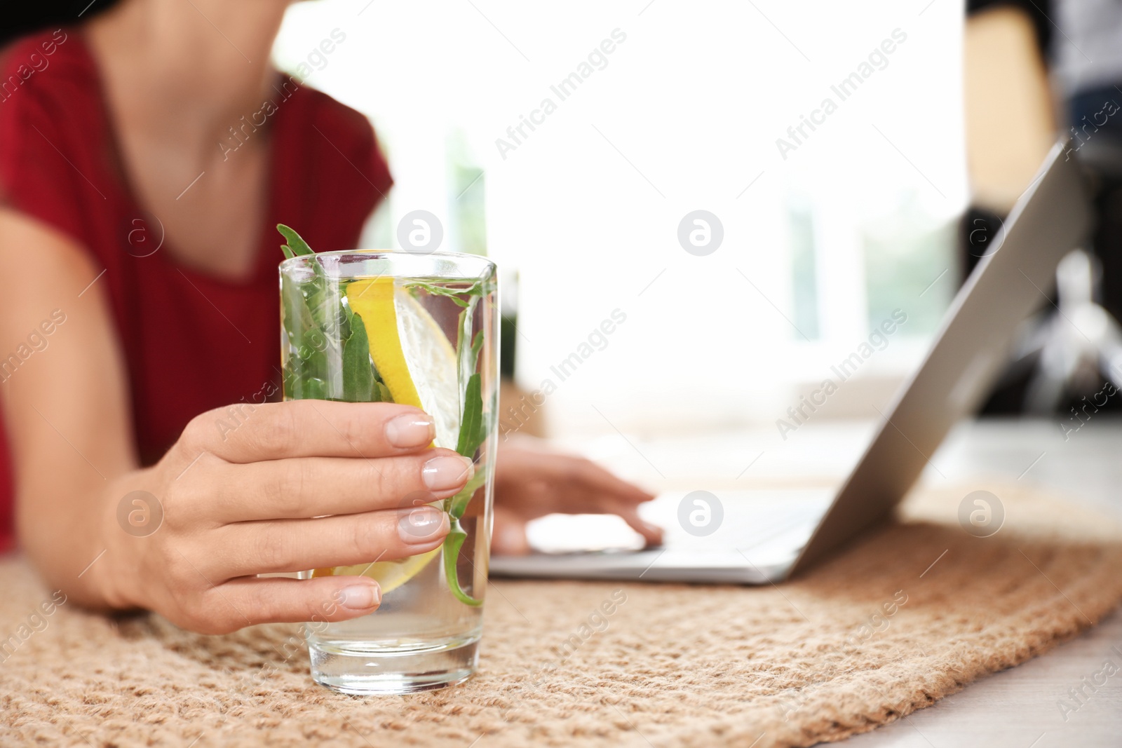 Photo of Young woman with glass of lemonade working on laptop at home, closeup. Refreshing drink