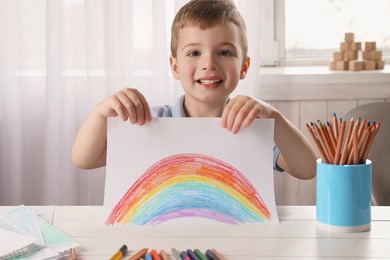 Photo of Child`s art. Cute little boy holding paper with drawingrainbow at white wooden table in room