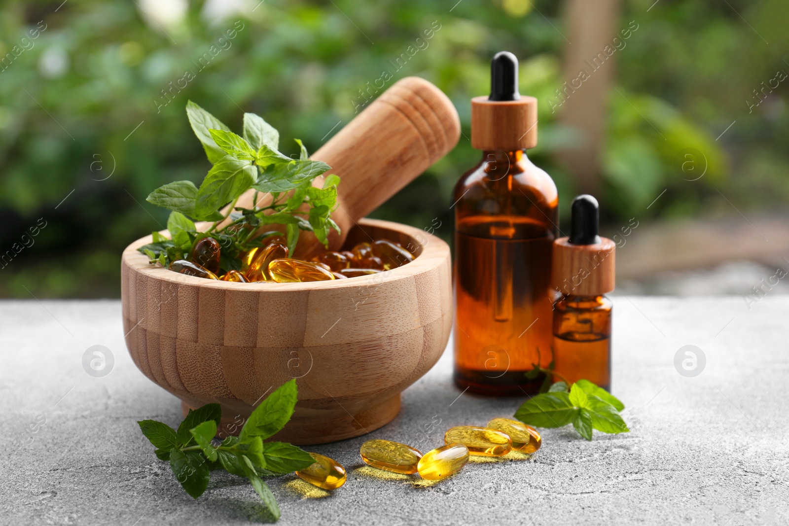Photo of Wooden mortar with fresh green herbs, extracts and capsules on light grey table outdoors