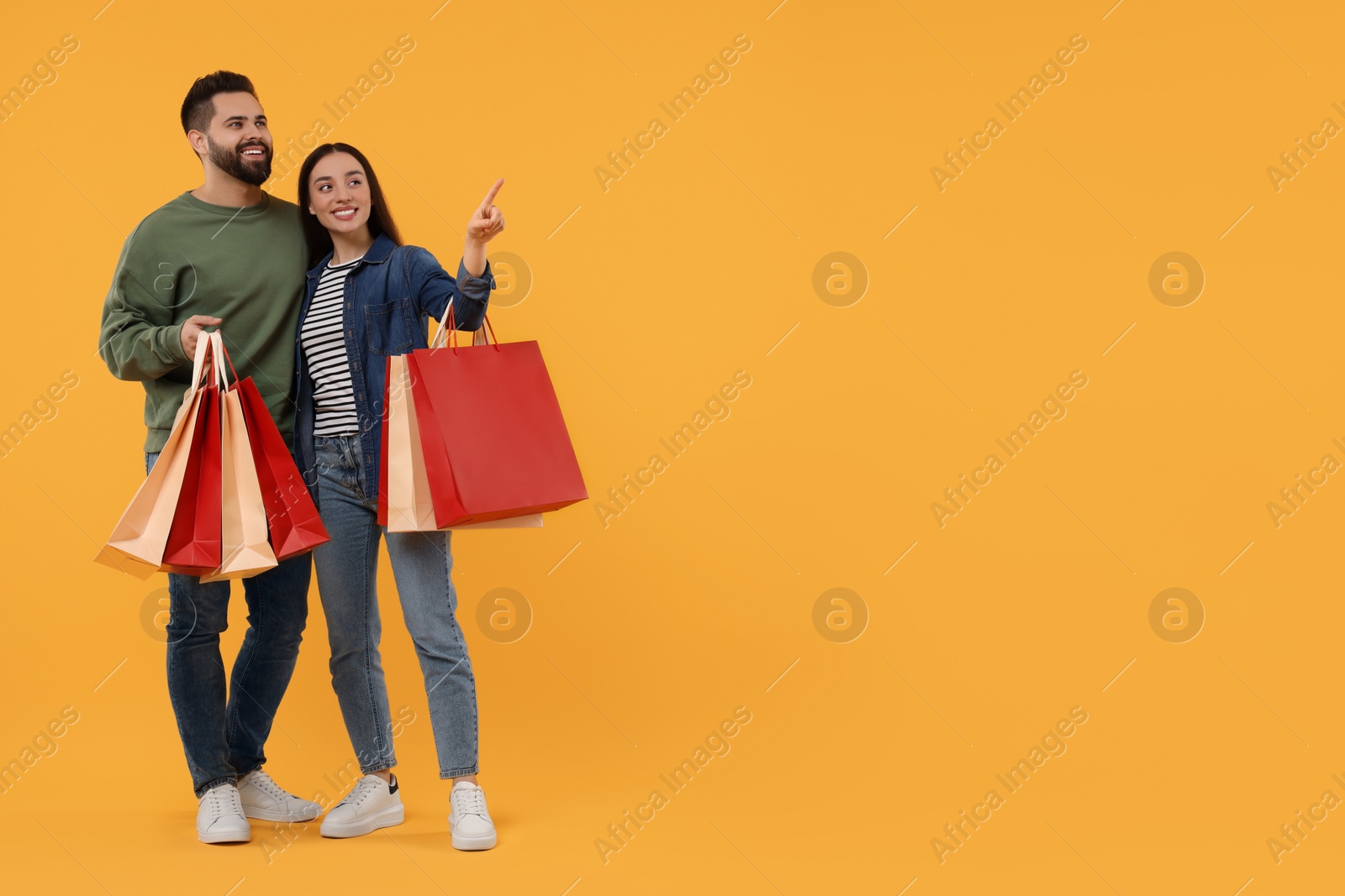 Photo of Happy couple with shopping bags looking at something on orange background. Space for text