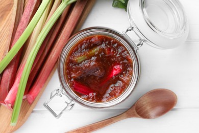 Jar of tasty rhubarb jam, stems and spoon on white wooden table, flat lay