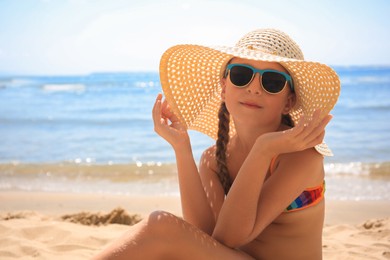 Photo of Little girl in stylish sunglasses and hat on sandy beach near sea