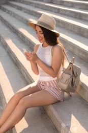 Photo of Beautiful young woman with stylish backpack and smartphone sitting on stairs outdoors