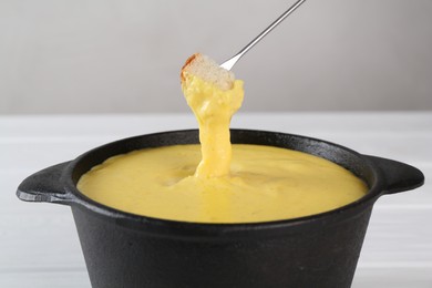 Dipping piece of bread into fondue pot with tasty melted cheese at white table, closeup