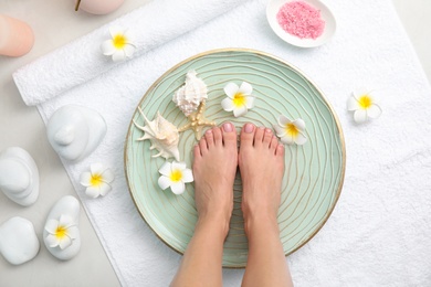 Photo of Woman soaking her feet in plate with water, flowers and seashells on white towel, top view. Spa treatment