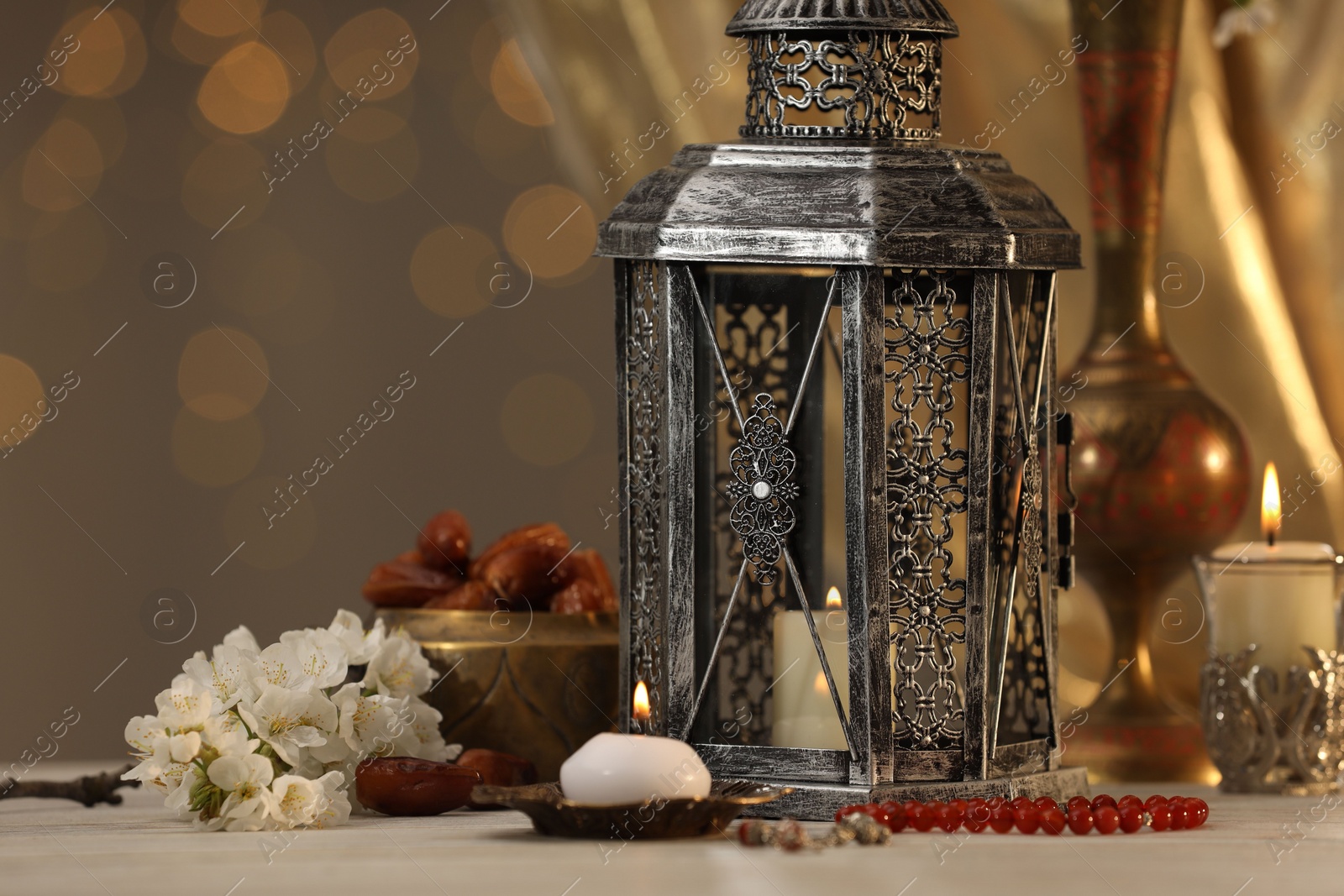 Photo of Arabic lantern, misbaha, candles, dates and flowers on table against blurred lights