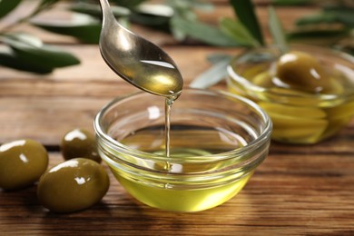 Spoon with cooking oil over bowl and olives on wooden table, closeup