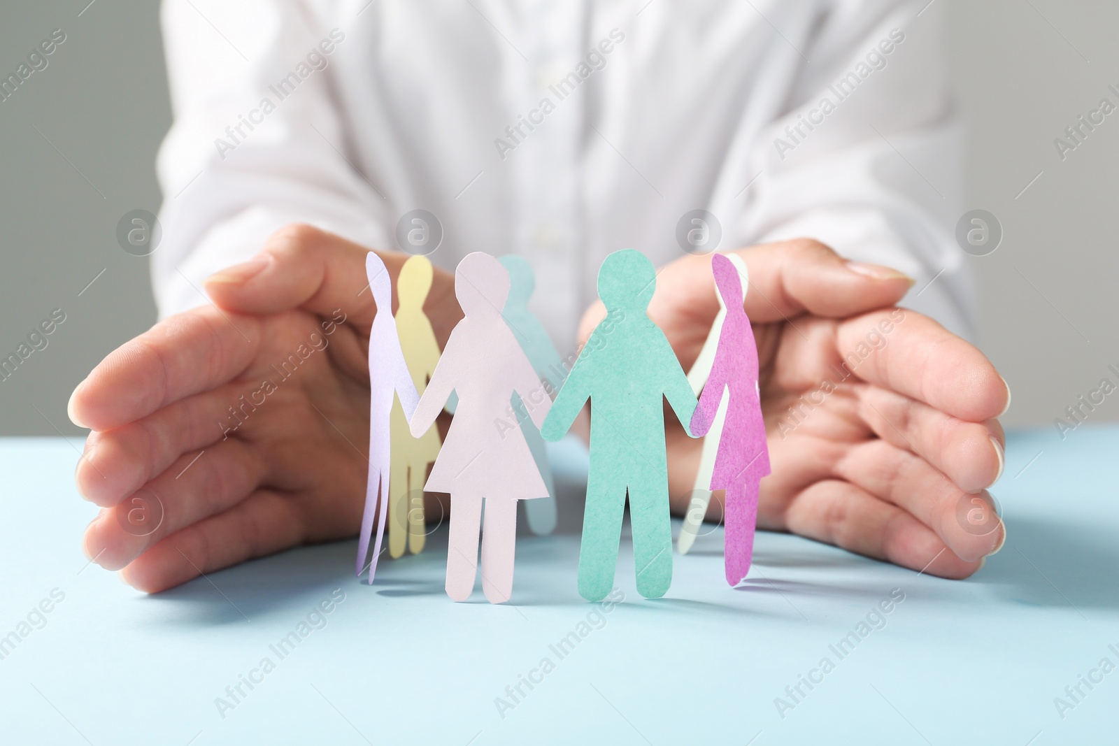 Photo of Woman protecting paper human figures on light blue table against grey background, closeup. Diversity and inclusion concept