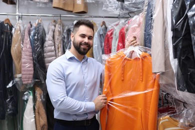 Photo of Dry-cleaning service. Happy worker holding hanger with hoodie in plastic bag near other clothes indoors