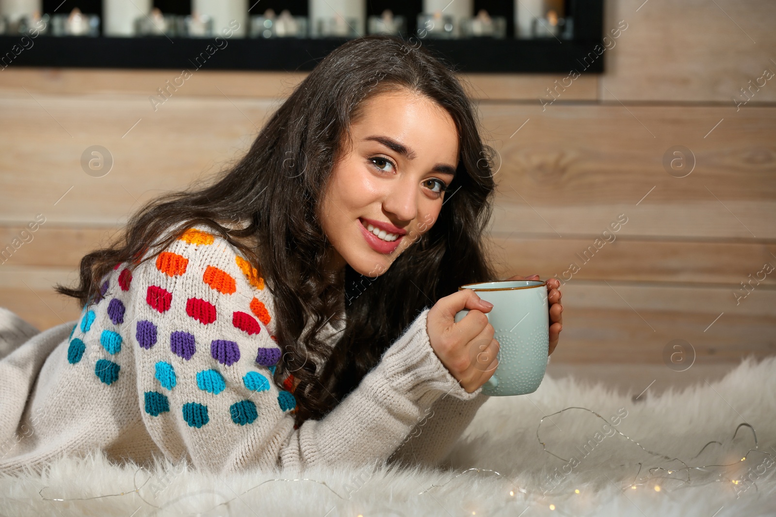 Photo of Young beautiful woman in warm sweater with cup of hot drink lying on rug at home
