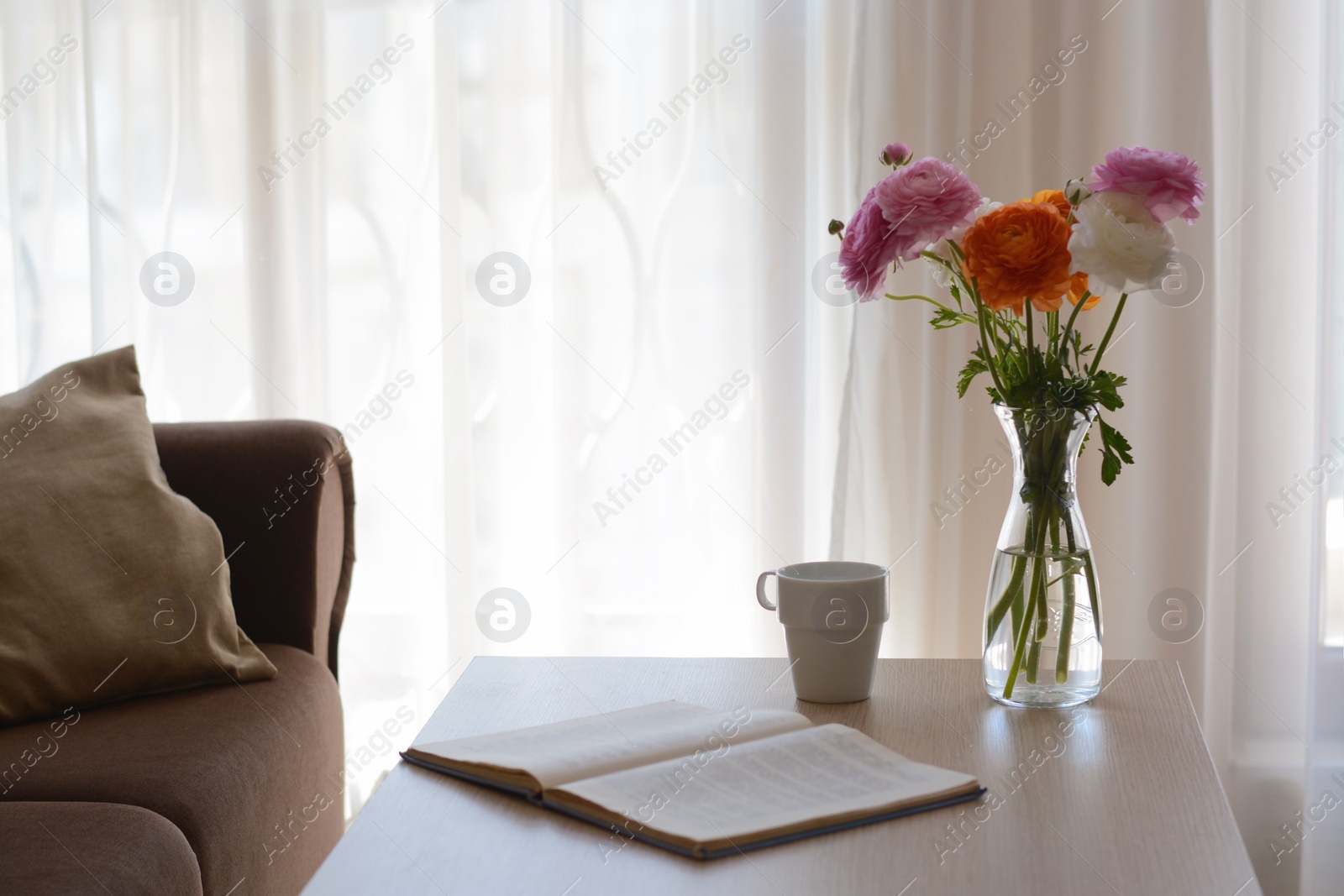 Photo of Bouquet of beautiful ranunculus flowers in vase, open book and cup on wooden table indoors. Space for text