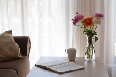 Bouquet of beautiful ranunculus flowers in vase, open book and cup on wooden table indoors. Space for text