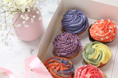 Photo of Different colorful cupcakes in box and flowers on white table, closeup