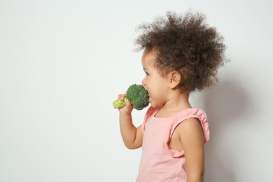 Photo of Cute African-American girl eating broccoli on white background. Space for text