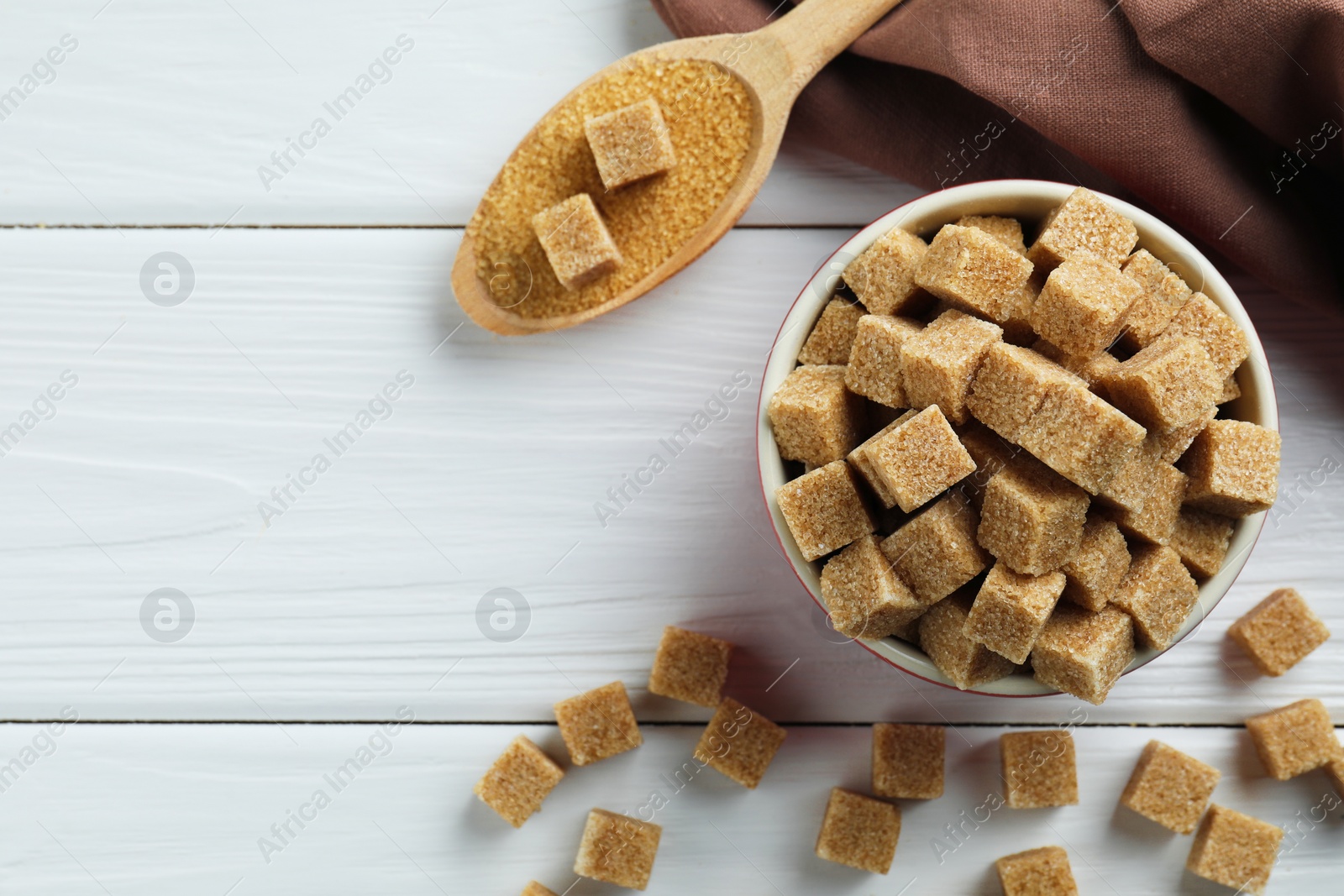 Photo of Bowl and spoon with brown sugar cubes on white wooden table, flat lay. Space for text