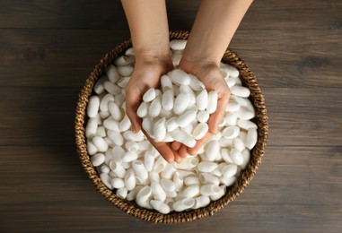 Photo of Woman holding white silk cocoons over bowl on wooden table, top view
