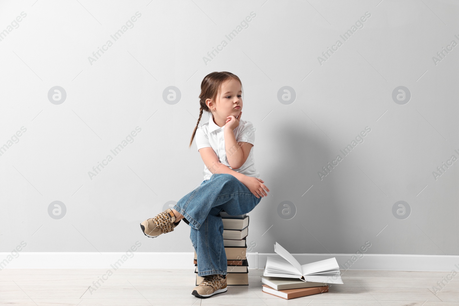 Photo of Cute little girl sitting on stack of books near light grey wall