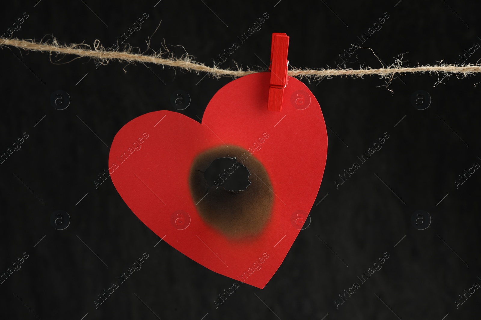 Photo of Red paper heart with burnt hole on rope against black background. Broken heart
