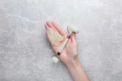 Woman holding gua sha tool and face roller on grey background, top view