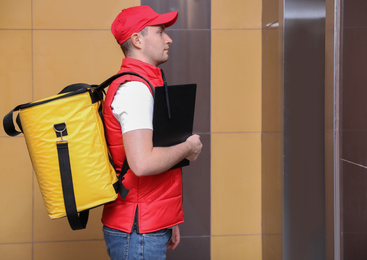 Male courier with thermo bag and clipboard waiting for elevator. Food delivery service