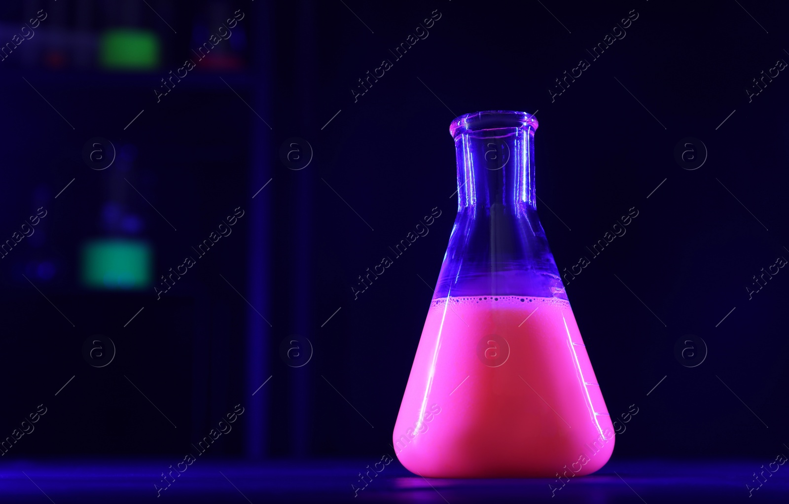 Photo of Laboratory flask with luminous liquid on table against dark background, space for text