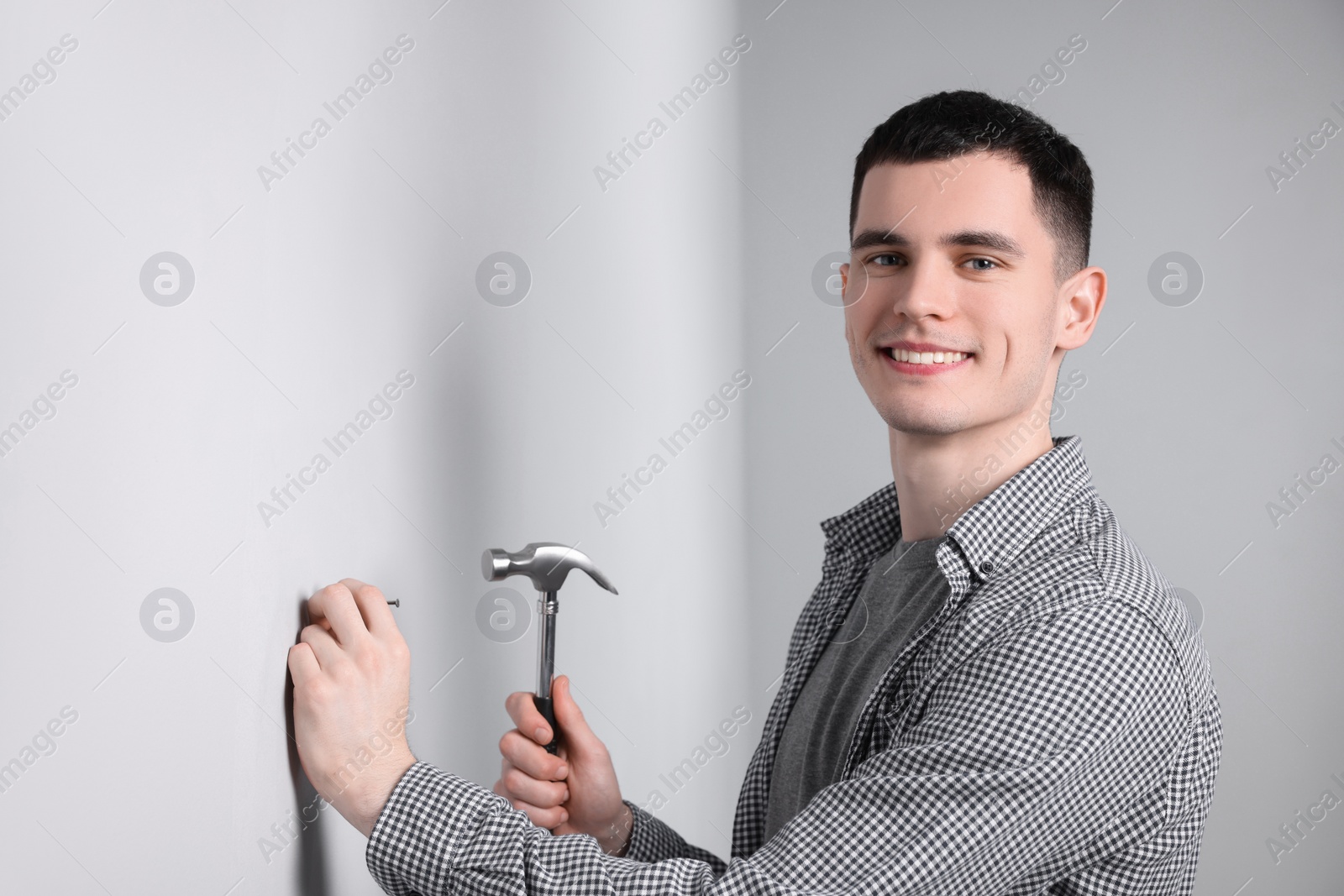 Photo of Young man hammering nail into white wall indoors