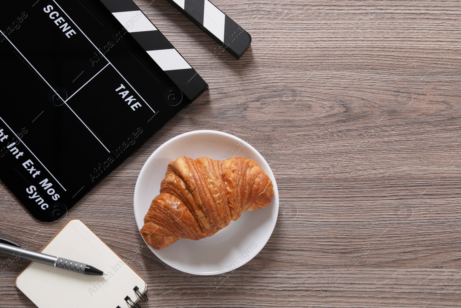 Photo of Movie clapper, croissant, notebook and pen on wooden table, flat lay. Space for text