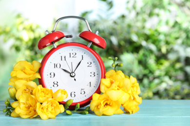 Red alarm clock and spring flowers on light blue wooden table against blurred greenery. Time change