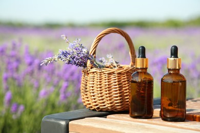 Photo of Bottles of essential oil and wicker bag with lavender flowers on wooden table in field outdoors, space for text