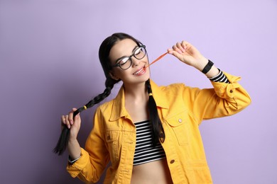 Fashionable young woman with braids chewing bubblegum on lilac background