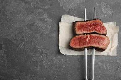 Photo of Carving fork with slices of delicious beef steak on grey table, top view. Space for text