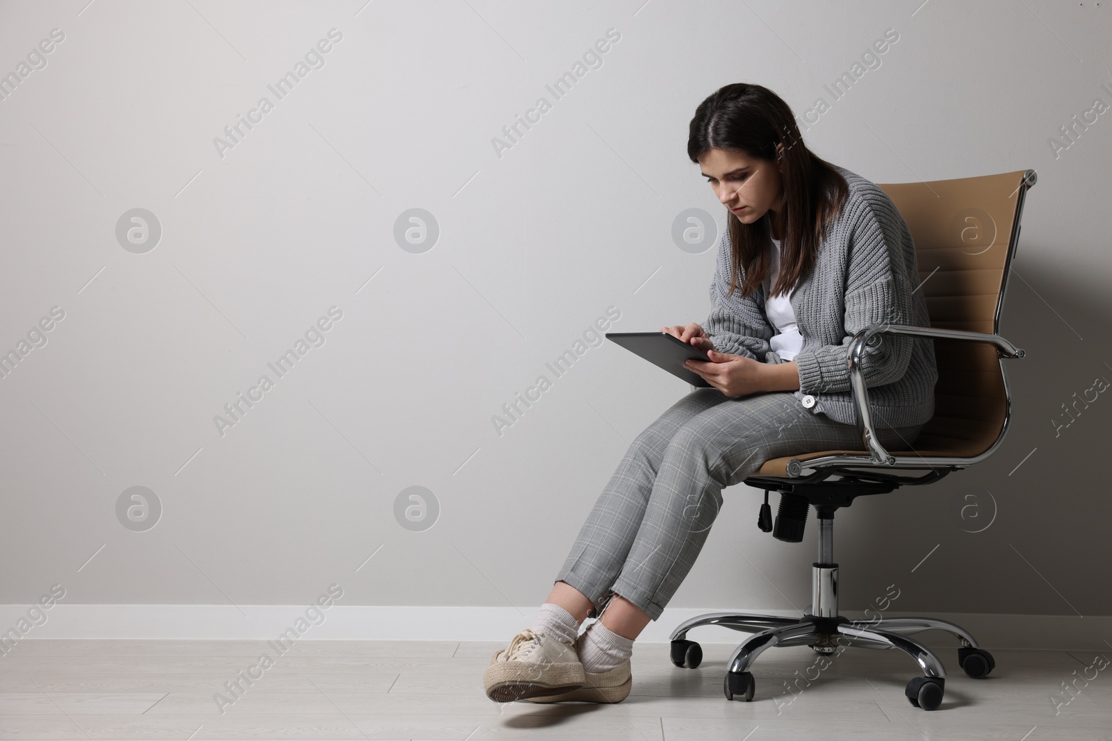 Photo of Young woman with bad posture using tablet while sitting on chair near grey wall. Space for text