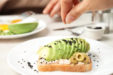 Woman adding black sesame to sandwich with avocado on plate, closeup