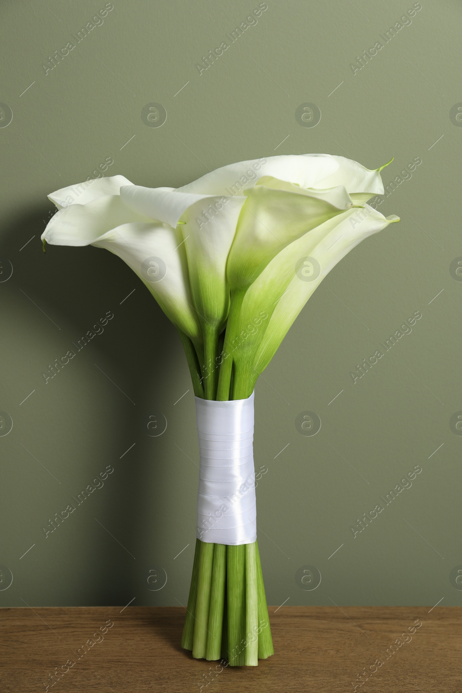 Photo of Beautiful calla lily flowers tied with ribbon on wooden table near olive wall
