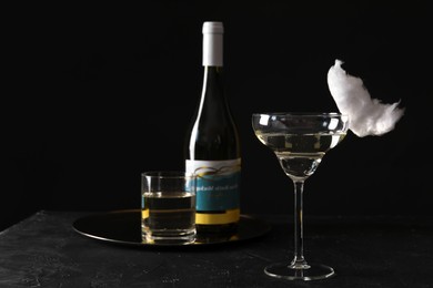 Photo of Cocktail with tasty cotton candy and bottle of alcohol drink on dark textured table against black background