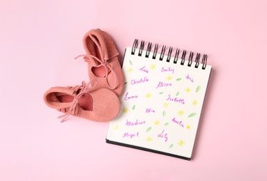 Photo of Notebook with different baby names and child's shoes on pink background, flat lay