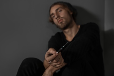 Young drug addict with syringe sitting near grey wall