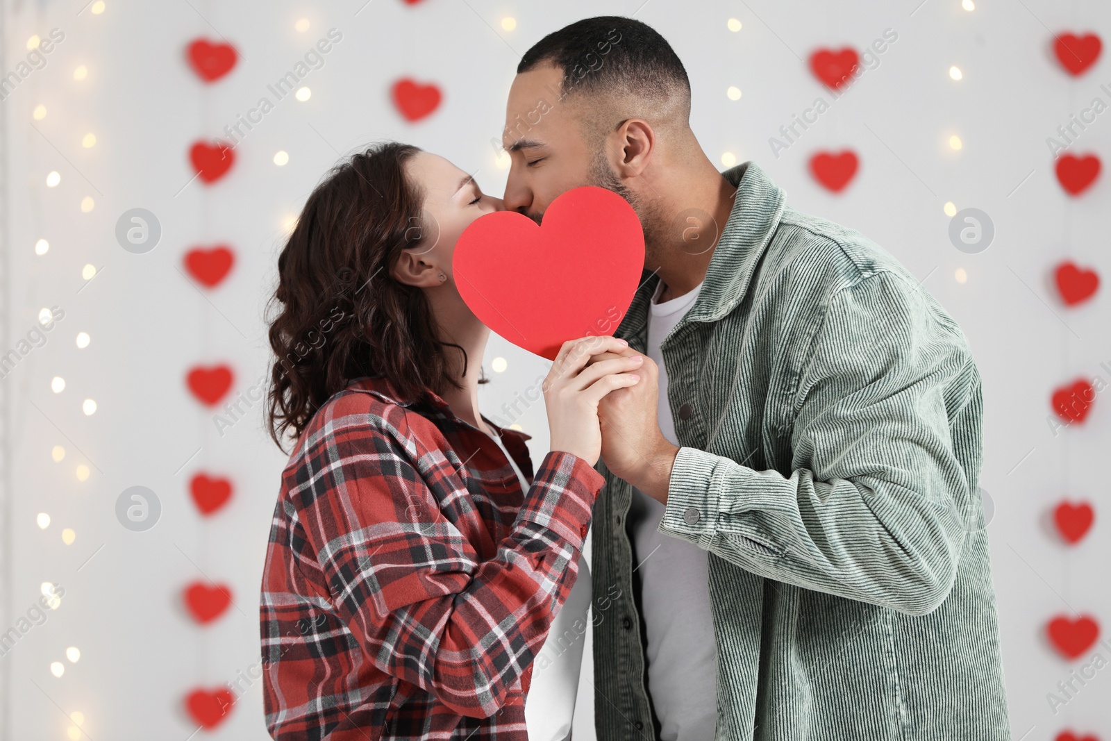 Photo of Lovely couple kissing behind red paper heart indoors. Valentine's day celebration