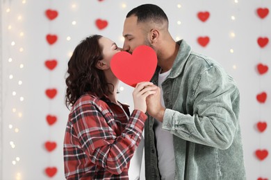 Photo of Lovely couple kissing behind red paper heart indoors. Valentine's day celebration