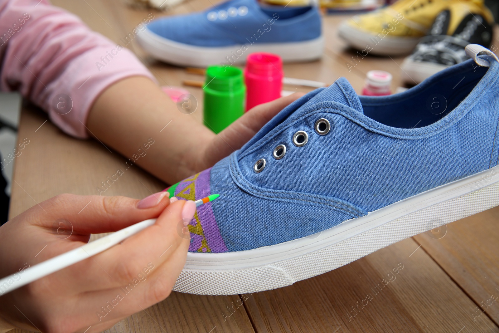 Photo of Woman painting on sneaker at wooden table, closeup. Customized shoes