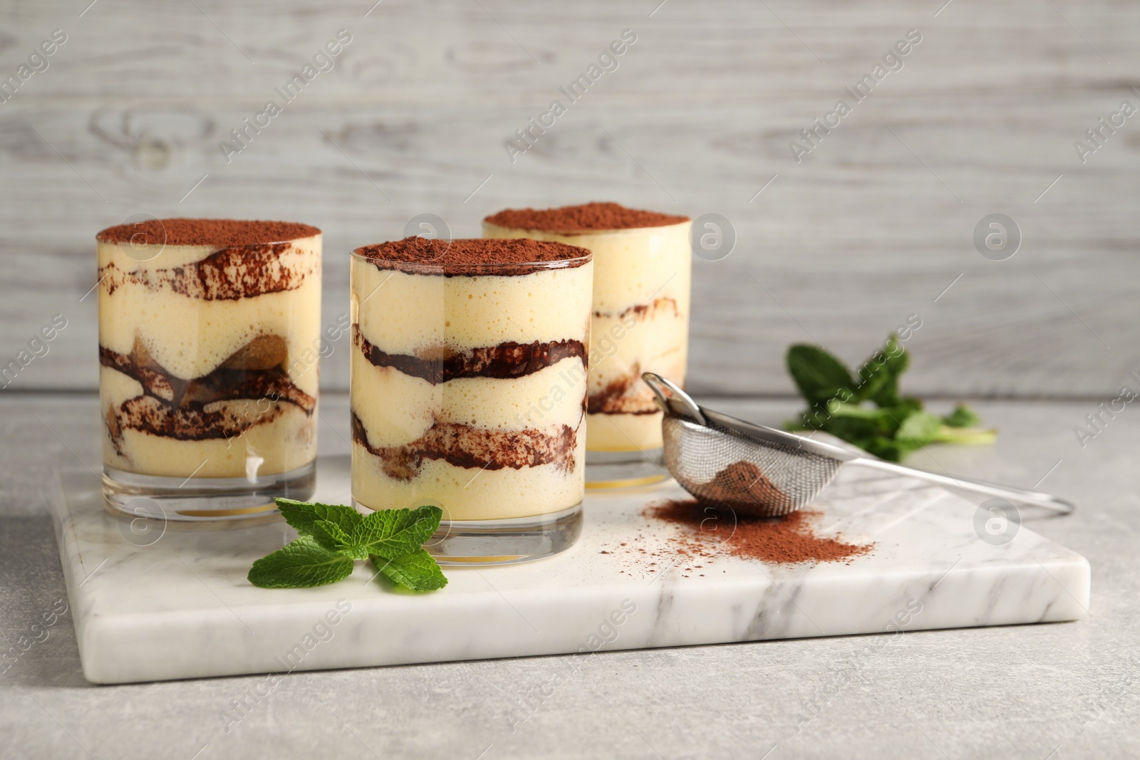 Photo of Delicious tiramisu in glasses, mint and sieve with cocoa powder on light textured table