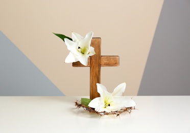 Photo of Wooden cross, crown of thorns and blossom lilies on table against color background