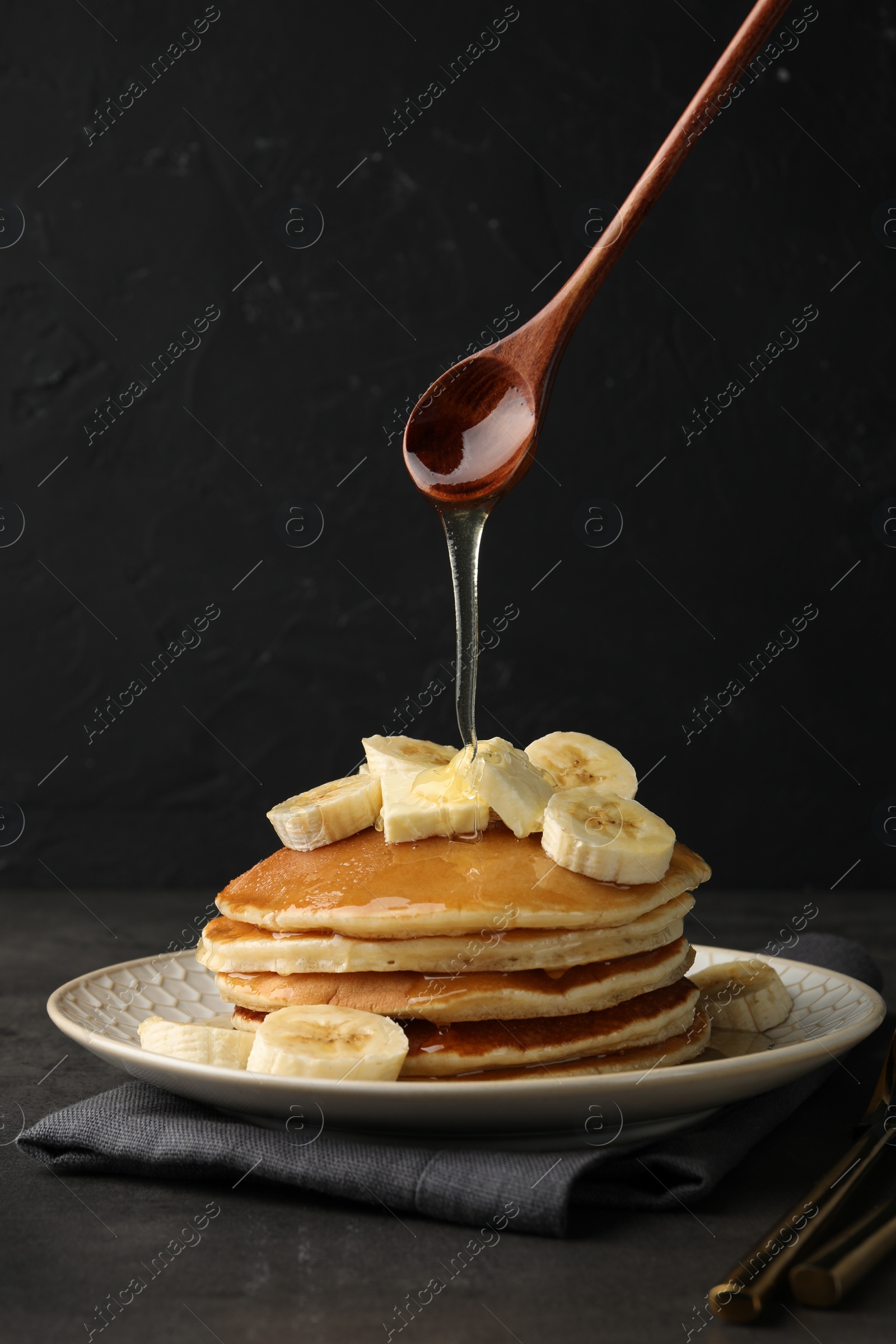 Photo of Pouring honey from spoon onto delicious pancakes with bananas and butter at dark table