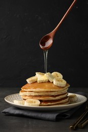 Photo of Pouring honey from spoon onto delicious pancakes with bananas and butter at dark table