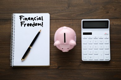 Image of Notebook with words Financial Freedom, piggy bank and calculator on wooden table, flat lay