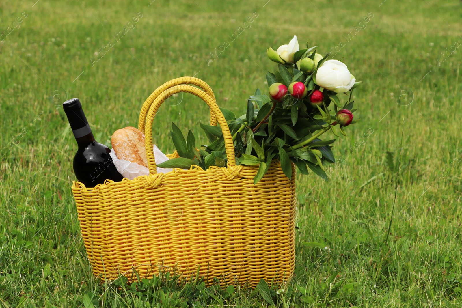 Photo of Yellow wicker bag with wine, bread and flowers on green grass outdoors. Picnic season