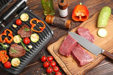 Photo of Electric grill with meat, spices, knife and vegetables on wooden table, flat lay