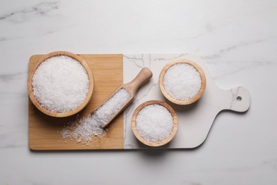 Wooden board with natural sea salt in scoop and bowls on white marble table, flat lay