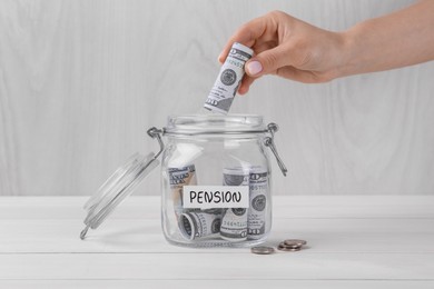 Photo of Pension concept. Woman putting dollar banknote into glass jar with money at white wooden table, closeup